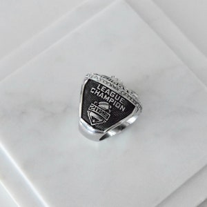 May include: A silver ring with a black and silver design. The ring features the words "League Champion" and the year "2023".
