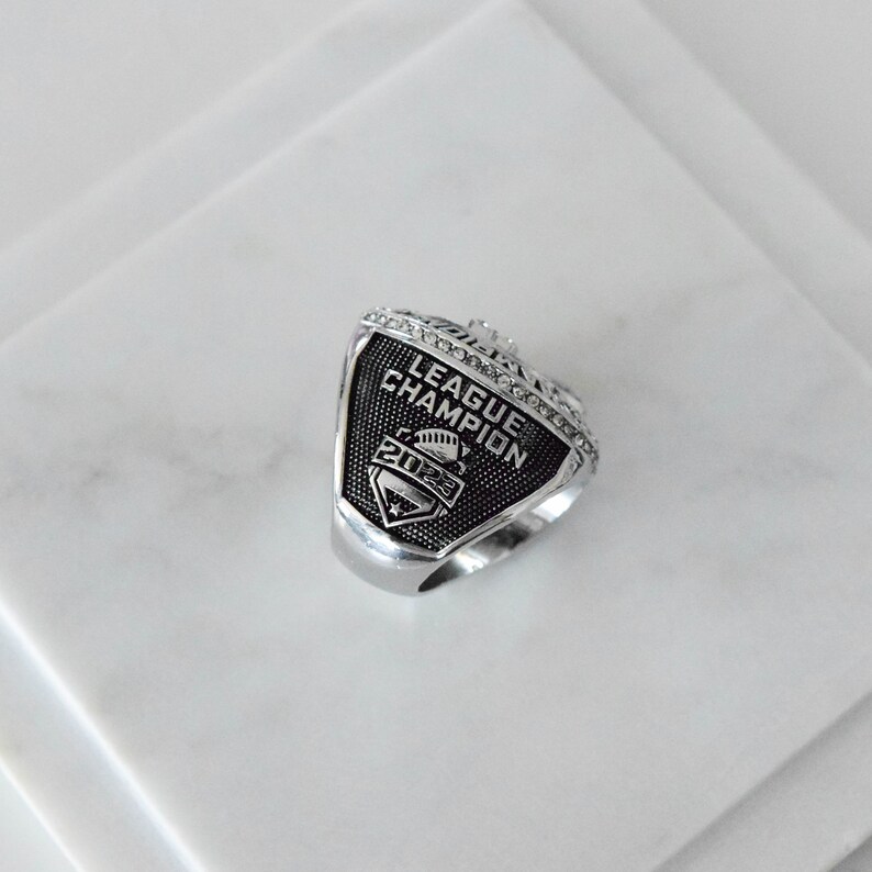May include: A silver ring with a black and silver design. The ring features the words "League Champion" and the year "2023".