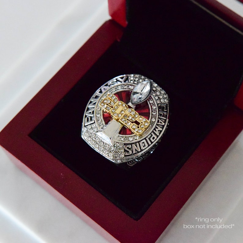 May include: A silver and gold championship ring with a red and white stone in the center. The ring says "Fantasy Champions" around the edge. The ring is in a red velvet box. *ring only, box not included*