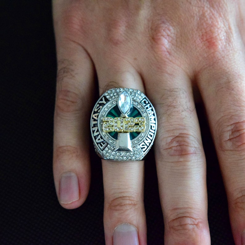 May include: A silver and green fantasy football championship ring with a clear stone and the text "Fantasy Champions 2022" around the ring.