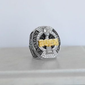 May include: A silver and gold fantasy football championship ring. The ring features the text "FANTASY CHAMPIONS" around the band and "2023" in gold lettering. The ring is encrusted with clear gemstones.