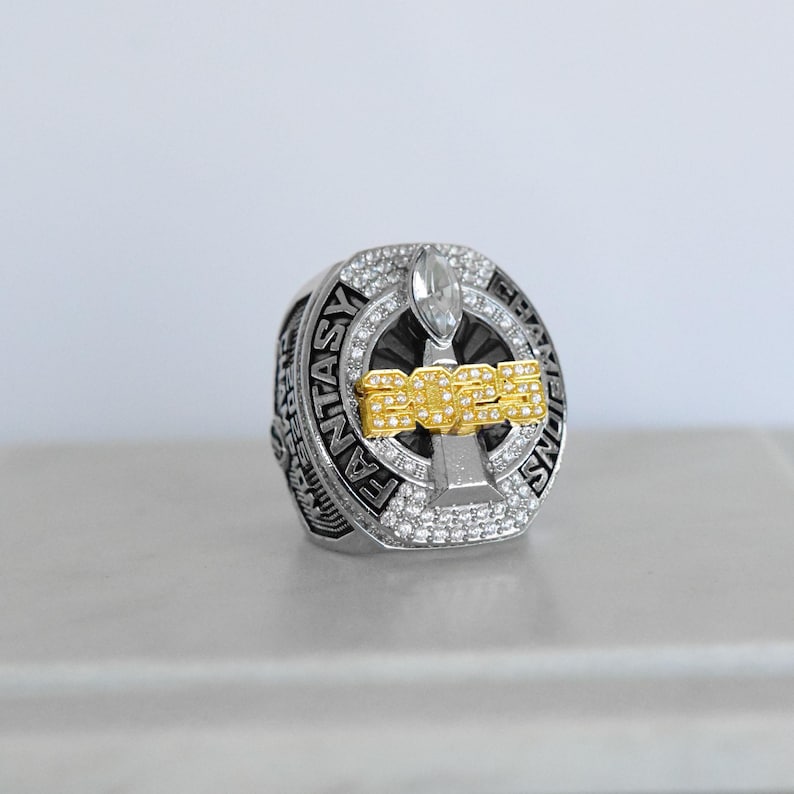 May include: A silver and gold fantasy football championship ring. The ring features the text "FANTASY CHAMPIONS" around the band and "2023" in gold lettering. The ring is encrusted with clear gemstones.