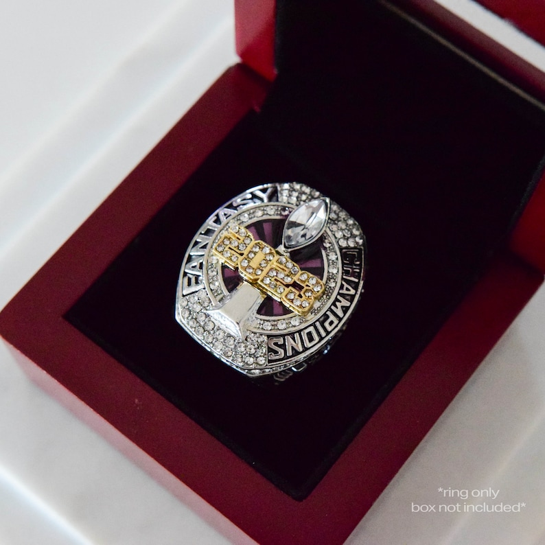 May include: A silver and gold championship ring with a purple stone and rhinestones. The ring features the words "Fantasy Champions" and a football trophy. *ring only, box not included*