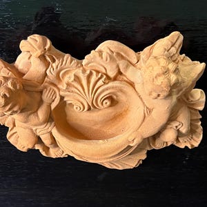 May include: A terracotta-colored cherub sculpture. Two cherubs flank a central bowl-shaped design, with detailed wings and floral accents. The sculpture is made of a light brown material.