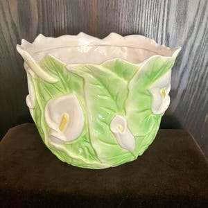 May include: A ceramic planter with a calla lily design. The planter features white calla lilies and green leaves against a light green background. The rim of the planter is white. The planter is a decorative item.