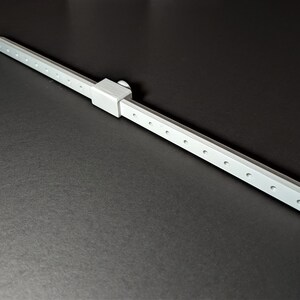 May include: A silver-colored, linear measuring tool with a sliding component and multiple evenly spaced holes. The tool is positioned diagonally against a dark background, suggesting a precision instrument.