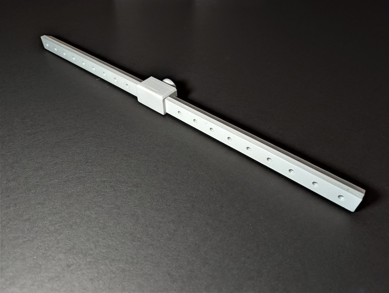 May include: A silver-colored, linear measuring tool with a sliding component and multiple evenly spaced holes. The tool is positioned diagonally against a dark background, suggesting a precision instrument.
