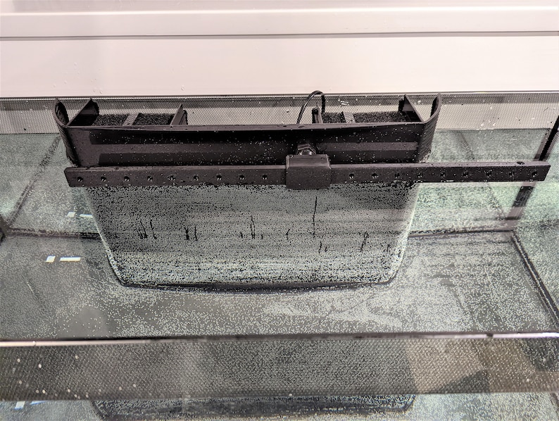 May include: A black aquarium filter with a rectangular design, featuring a filtration compartment and an adjustable arm. The filter is submerged in water, with bubbles visible, suggesting active aeration. The filter is designed to fit on the edge of the aquarium.