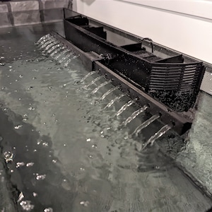 May include: A black aquarium filter system with water cascading down multiple spouts into a clear tank. The water is bubbling, and the filter is positioned above the water level. The tank has a mesh side.