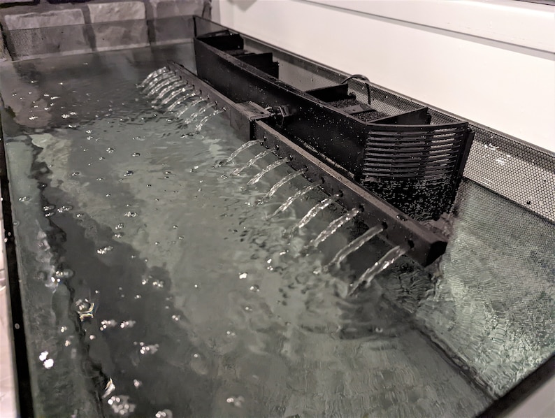 May include: A black aquarium filter system with water cascading down multiple spouts into a clear tank. The water is bubbling, and the filter is positioned above the water level. The tank has a mesh side.