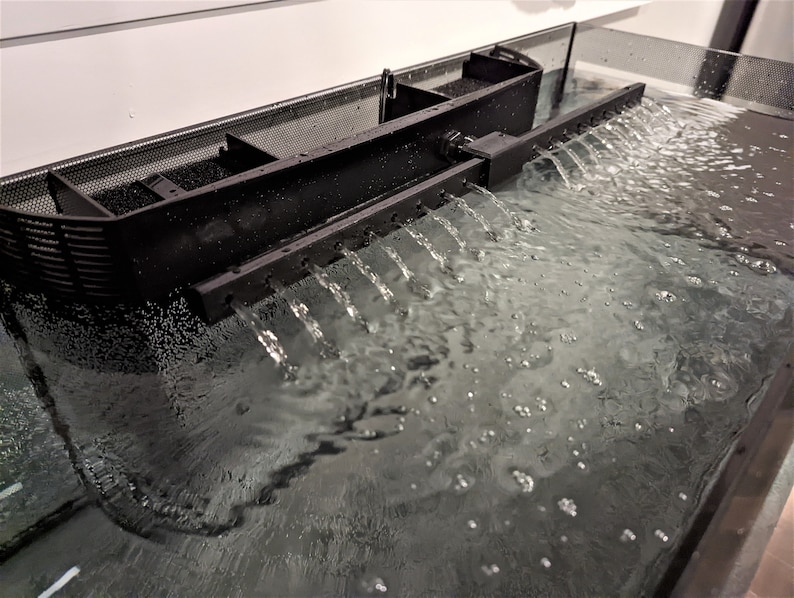May include: A black aquarium filter with water flowing through it. The filter has a rectangular shape with a mesh top and multiple outlets. Water cascades from the outlets into the aquarium, creating a small waterfall effect. The water is clear and the filter is positioned near the top of the tank.