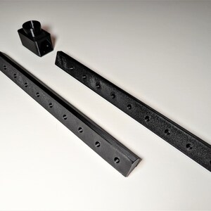 May include: Black 3D-printed components, including two long, rectangular bars with evenly spaced holes and a small, angled piece. The bars have a textured surface. The pieces are arranged on a white surface.