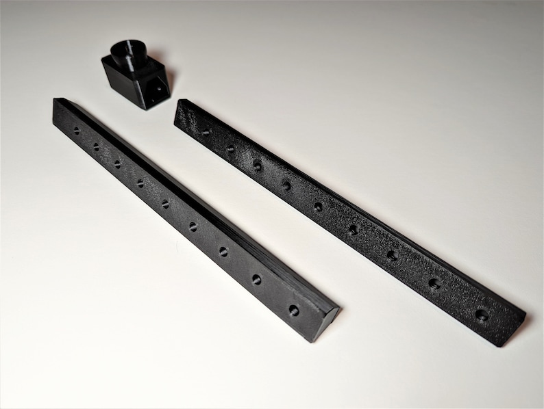 May include: Black 3D-printed components, including two long, rectangular bars with evenly spaced holes and a small, angled piece. The bars have a textured surface. The pieces are arranged on a white surface.