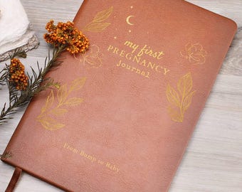 May include: A brown leather-textured journal with gold foil lettering that reads "my first PREGNANCY journal" and "From Bump to Baby," decorated with gold line-art illustrations of leaves, flowers, a crescent moon, and stars.