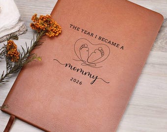 May include: A tan leather-like journal featuring the black printed text THE YEAR I BECAME A mommy 2026 and a line-art illustration of baby feet inside a heart shape. A small sprig of orange berries is positioned on the left side of the cover.