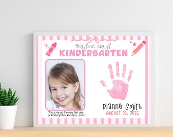 May include: A rectangular sign with a pink and white striped border featuring the text My first day of KINDERGARTEN, a photograph of a child, a pink handprint, the name Dianne Smith, the date AUGUST 10, 2026, and a crayon and pencil graphic.