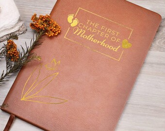 May include: A brown leather-textured journal with a gold foil design featuring the text THE FIRST CHAPTER OF MOTHERHOOD, a pair of baby footprints, and a heart. A gold line-art floral illustration is below the text, and a ribbon bookmark is attached.
