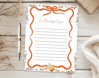 May include: A white rectangular stationery sheet featuring an orange ribbon border with a bow at the top and a small pumpkin at the bottom. The text "I'm thankful for" is printed in orange above twelve horizontal lines for writing, accompanied by a pen.