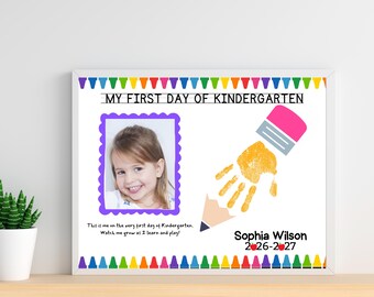 May include: A framed rectangular sign titled MY FIRST DAY OF KINDERGARTEN with a colorful crayon border. It features a photo of a child in a purple frame, a yellow handprint shaped like a pencil, and the text Sophia Wilson 2026-2027.