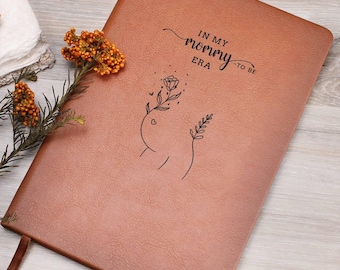 May include: A tan leather-like journal featuring the black printed text IN MY MOMMY ERA and a line art illustration of a pregnant torso with a flower and a small heart. A sprig of dried orange flowers rests on the left side of the notebook cover.