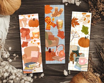 May include: Three rectangular bookmarks featuring autumn-themed illustrations. Designs include pumpkins, cozy armchairs, stacks of books, scarves, coffee cups, and falling leaves. The bookmarks have white backgrounds with grid or stripe patterns.