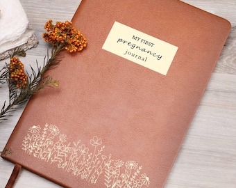 May include: A brown leather-textured journal with a rectangular label reading MY FIRST pregnancy journal in black text. The cover features a gold-colored line art illustration of wildflowers along the bottom edge and a matching brown ribbon bookmark.