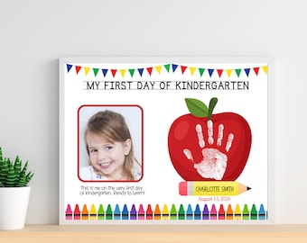 May include: A framed print titled MY FIRST DAY OF KINDERGARTEN with colorful bunting and crayon borders. It features a photo of a child, a red apple graphic with a white handprint, and the text Charlotte Smith and August 15, 2026.