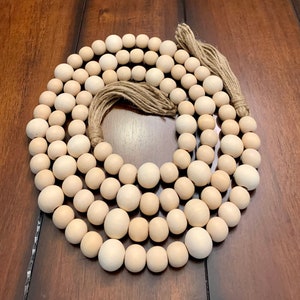 May include: A long strand of natural wood beads, approximately 50 beads in total, with a jute twine tassel at the end. The beads are a light beige color and are arranged in a circular pattern on a wooden surface.