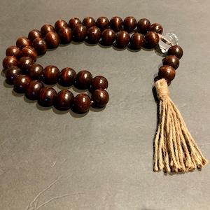 May include: A strand of dark brown wooden beads with a clear crystal accent and a natural fiber tassel. The beads are round and evenly spaced, creating a simple, elegant design.