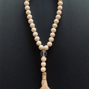 May include: A decorative garland featuring a continuous loop of smooth, light-colored wooden beads. A single clear, spherical glass bead is positioned near the bottom, followed by three additional wooden beads and a frayed jute fiber tassel.