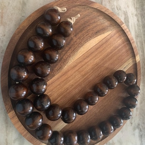 May include: A wooden beaded garland with dark brown, round beads, displayed on a round wooden tray. The beads are strung on a natural twine. The tray has a two-tone wood grain pattern.
