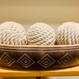 May include: Three white rope balls in a dark brown wooden bowl with a geometric design. The bowl is oval shaped and has a carved pattern of diamonds.