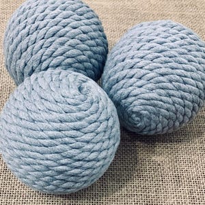May include: Three light blue, spherical decorative objects made of tightly wound rope. The objects are arranged in a cluster, with the texture of the rope clearly visible. The background is a textured, neutral-toned fabric.
