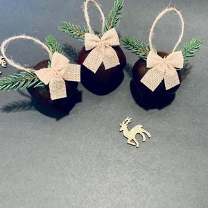 May include: Three dark brown Christmas ornaments with burlap bows and faux evergreen sprigs. Each ornament has a twine loop for hanging. Small wooden reindeer decorations are scattered around the ornaments.
