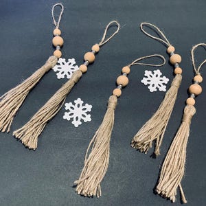 May include: Five jute tassels with wooden beads and twine loops for hanging, interspersed with white snowflake cutouts. The tassels are a natural beige color, and the beads are light brown.