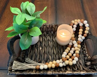 May include: A woven tray with handles holds a small potted plant with green leaves, a lit candle in a glass holder, and a decorative wooden bead garland. The tray is dark brown and sits on a wooden surface.