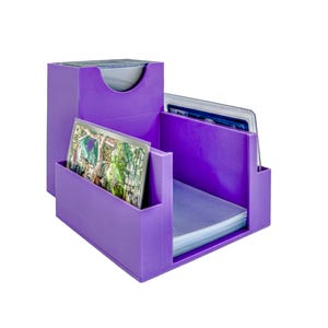 Puede incluir: Una caja de almacenamiento de tarjetas morada con múltiples compartimentos. La caja tiene forma rectangular con una abertura curva en la parte superior. Varias tarjetas son visibles en los compartimentos. Diseñada para organizar y guardar tarjetas.
