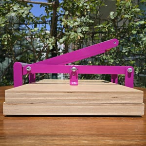 May include: A magenta-colored book press with a wooden base. The press features a lever arm and a screw mechanism for applying pressure. The wooden base is made of layered plywood. The press is designed for crafting or bookbinding.