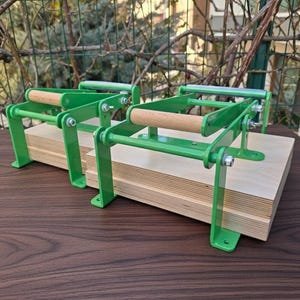 May include: Two green metal tortilla presses with wooden handles and a light-colored wooden base. The presses are designed with a lever mechanism and are set on a dark wooden surface.