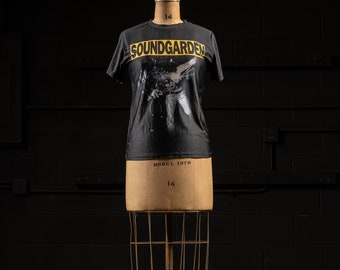 May include: A black short-sleeve t-shirt featuring the word SOUNDGARDEN in yellow capital letters across the chest, positioned above a black and white graphic print of a person in motion. The shirt is displayed on a vintage dress form mannequin.