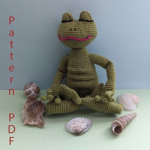 May include: A green crocheted frog sitting in a yoga pose with its eyes closed. The frog is surrounded by seashells on a light green surface. The text "Frog Yogi" is at the top of the image and the text "Pattern PDF from Inna" is at the bottom of the image.