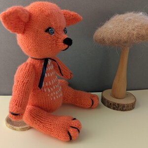 Könnte beinhalten: Ein gestricktes orangefarbenes Fuchsspielzeug mit schwarzen Augen und einer schwarzen Nase, das auf einem Holzsockel sitzt. Der Fuchs trägt ein schwarzes Band um den Hals. Der Text "Knitted fox pattern" und "From Inna" ist auf dem Bild zu sehen.