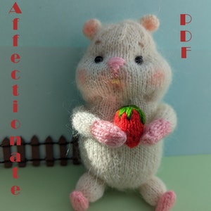 May include: Knitted white hamster holding a red strawberry. The hamster is sitting on a green surface with a wooden fence in the background. The text "PATTERN PDF" is in red on the right side of the image. The text "AFFECTONATE HAMSTER" is in red on the left side of the image.