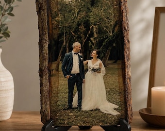 May include: A rectangular wooden plaque with natural bark edges featuring a printed photograph of a couple in wedding attire standing in a wooded area. The plaque is supported by a black metal stand, displaying a clear image of the pair.