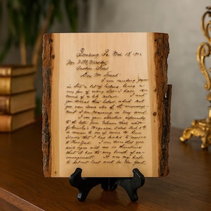 Puede incluir: Placa de madera con una carta manuscrita, exhibida sobre un soporte negro. La carta está en cursiva y fechada el 18 de marzo de 1922. La placa tiene un borde de corteza natural, añadiendo un toque rústico.