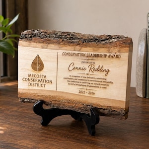 Puede incluir: Una placa rectangular de madera con bordes de corteza natural, apoyada sobre un soporte negro. La placa presenta el texto grabado "CONSERVATION LEADERSHIP AWARD", "Mecosta Conservation District", "Connie Redding" y el rango de fechas "2022-2026".