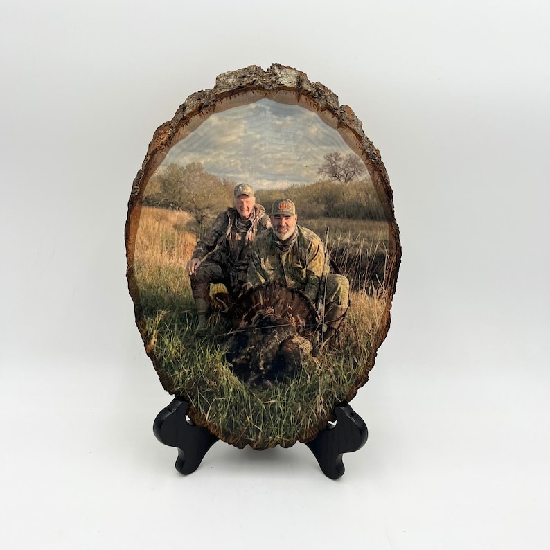 May include: A vertical oval-shaped wood slice plaque featuring a printed photograph of two individuals in camouflage clothing posing with a wild turkey in a grassy field. The natural bark edges are visible, and the plaque rests on a black stand.