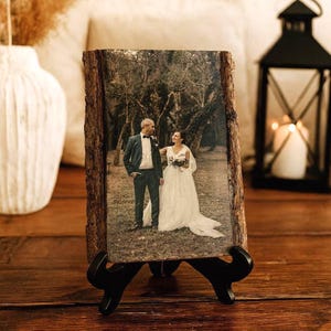 Peut inclure: Une plaque en bois rectangulaire avec des bords en écorce naturelle présentant une photographie imprimée d'un couple en tenue de mariage dans une zone boisée. La plaque est soutenue par un support en plastique noir sur une surface en bois.
