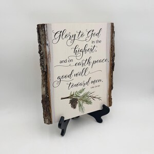May include: A wooden sign with a rustic bark edge features a Christmas-themed quote: "Glory to God in the highest, and on earth peace, good will toward men. Luke 2:14 KJV". The sign is decorated with a green pine branch and a pine cone.