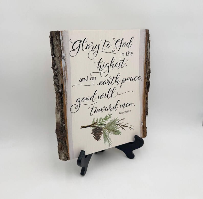 May include: A wooden sign with a rustic bark edge features a Christmas-themed quote: "Glory to God in the highest, and on earth peace, good will toward men. Luke 2:14 KJV". The sign is decorated with a green pine branch and a pine cone.